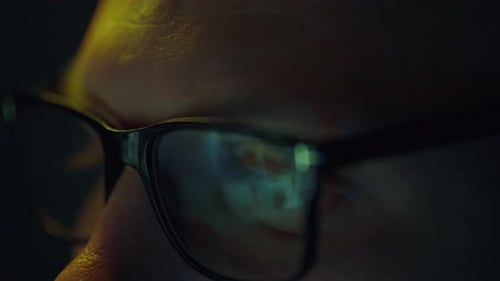 A programmer's eyes are locked in close-up on a bright computer monitor as he works remotely from