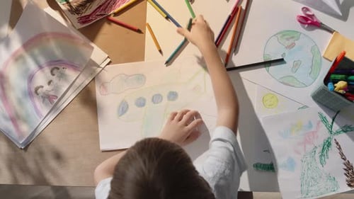Child Drawing Airplane at Table Covered in Art