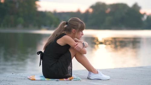 Woman Sitting by Lake at Sunset Hugging Knees