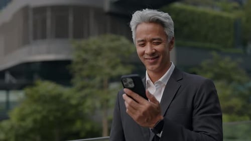Portrait of Mature Asian Man Manager Smiles and Looks at Smartphone in Park