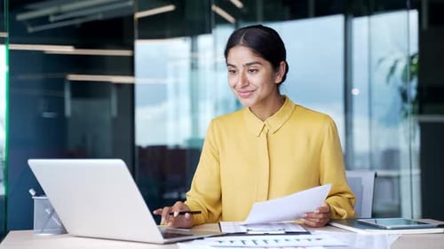 Happy young businesswoman sitting at desk in modern office looking at computer and financial