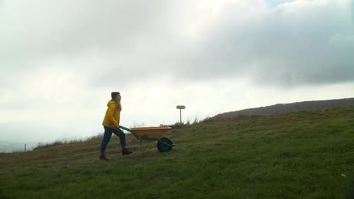 Young Woman Push Wheel Cart Up Green Hill