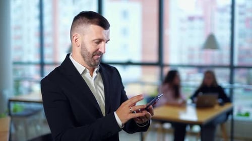 Businessman standing in office and holding phone. Entrepreneur messaging to somebody.