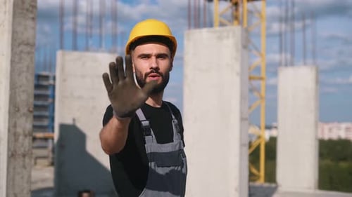 Showing stop gesture by hand. Young engineer in hard hat and uniform is on the construction site