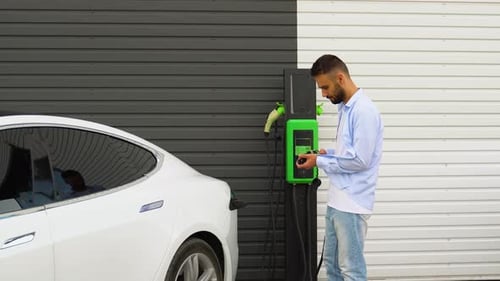 Stylish Man Plugging in Charging Cable to Electric Vehicle and Charges Batteries at Station