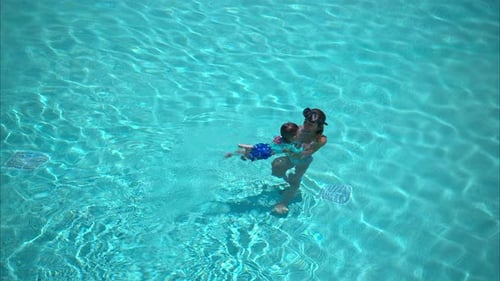 Happy latin hispanic mother wearing a mask teaching her son how to swim on a big pool on a bright su