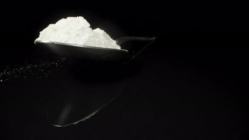 A spoon with a heap of white powder on a black background, illuminated by flickering light, close-up