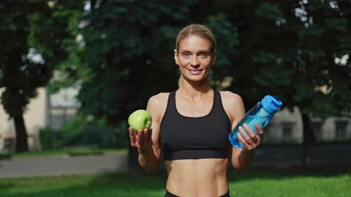Portrait of Sporty Woman Standing at Park with Green Apple and Bottle of Water