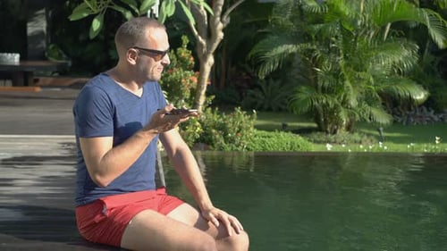 Attractive young man chatting on cellphone by the swimming pool at a tropical resort