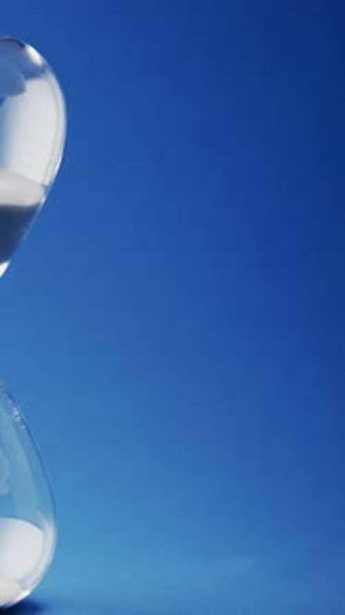 Vertical video of close up of hourglass with white sand pouring with copy space on blue background