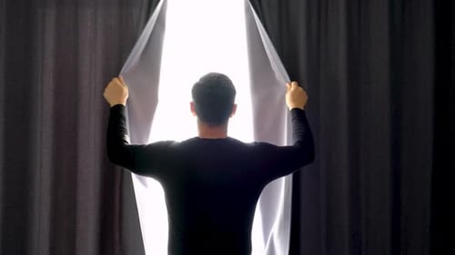 Young man in black t-shirt looks out window and opens the curtains, rear view.