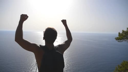 Unrecognizable Hiker with Backpack Standing on Edge of Mountain and Victoriously Raised Hands Male