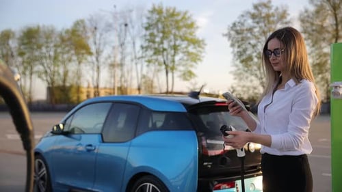 Kaukasische junge fröhliche Frau ruft in der Stadt mit dem Smartphone an und wartet darauf, dass ihr Elektroauto aufgeladen wird