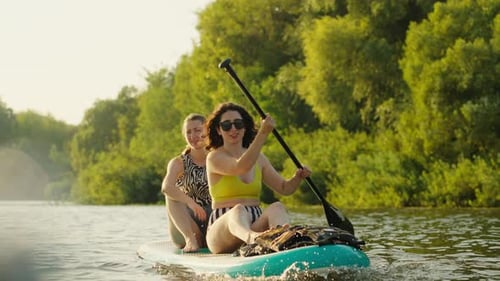 Paddle Boarding Sport In River In Summertime Two Beautiful Women Floating On One Board