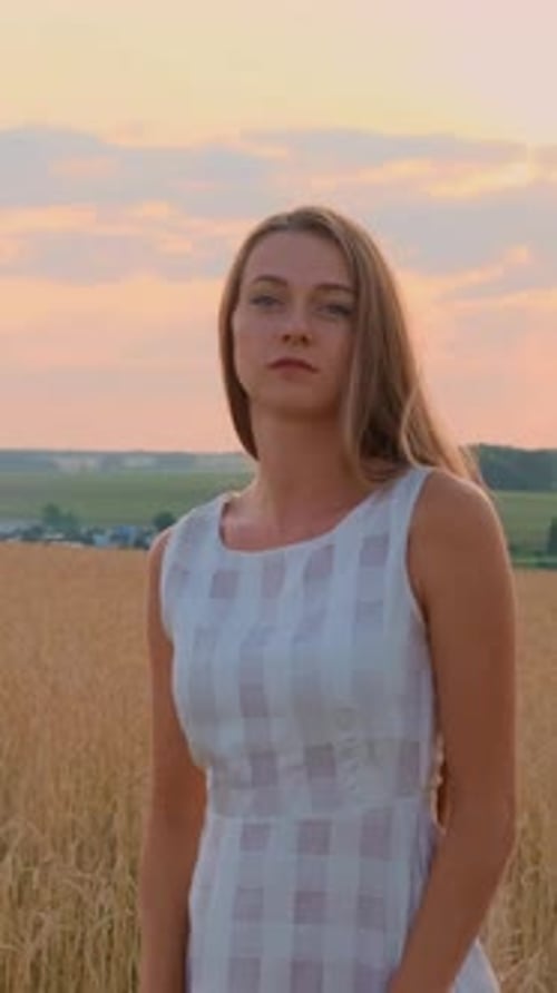 Young Woman in a Light Dress Poses Gracefully in a Golden Field During Sunset with Soft Clouds