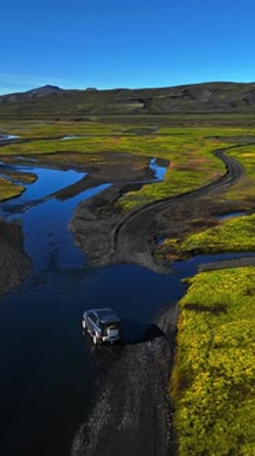 Drone aerial of a 4x4 SUV crossing a shallow river beside a gravel road in vivid green moss and