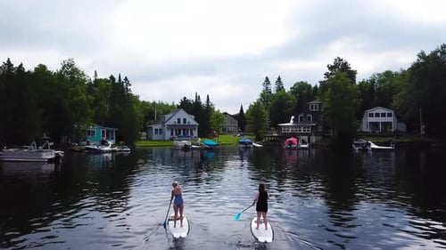 Two women paddle towards the lake houses that are on the waterfront and the shoreline of the lake. T