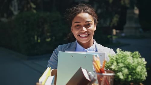 Smiling Businesswoman Carrying Office Supplies Outdoors in Bright Sunlight