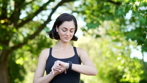 A young cute female runner stands in an urban city park and looking smart watch. Sportswoman using