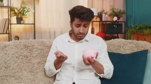 Indian Man Feeling Frustrated Looking at Empty Piggy Bank with Few Coins Facing Financial Crisis
