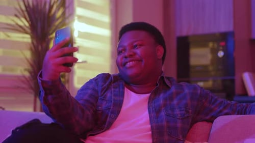 Funny Smiling Young African American Man Typing Message and Looking at Smartphone Screen Sitting on