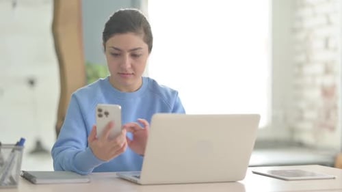 Young Woman Uses Phone and Laptop at Desk