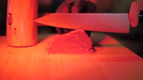 Unrecognizable chef cutting slice of raw beef meat for steak, close up with red light tone