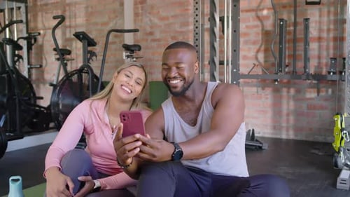 Casal diverso rindo enquanto homem estende o smartphone com capa rosa, mostrando fotos na academia em casa
