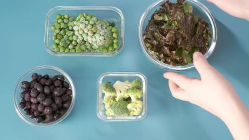Main féminine tenant une boîte en verre avec salade verte sur fond bleu, vue de dessus.
