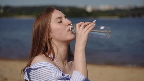 A Girl Drinks Water From a Plastic Bottle While Sitting on a Sandy Beach Near a River