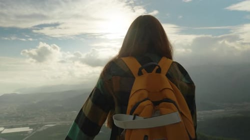 At the mountain summit, a woman watches the sunset, a backpack behind her, and her hair billowing.