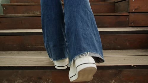 Young Woman Wearing Blue Jeans and White Sneakers Walking on Wooden Stairs at Sunny Day Back View