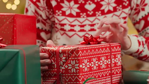 Woman Holding Wrapped Christmas Gift on Table with Wrapping Paper and Ribbon