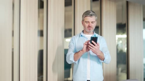 Happy mature gray-haired bearded business man in casual clothes using cellphone standing in modern
