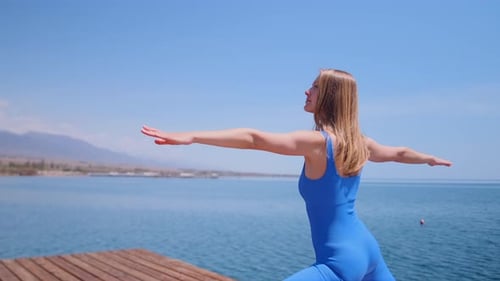 Frau führt Warrior II Yoga-Pose auf, Seitenansicht, während die Kamera auf einem Holzsteg am Meer im Freien nach links schwenkt