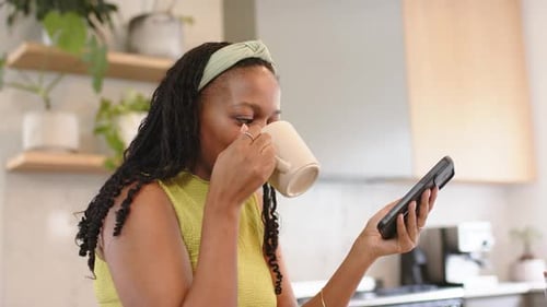 Raising mug, African American woman sipping coffee scrolling phone in kitchen reacting to text