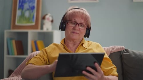 Elderly Woman with Headphones Works Remotely on a Tablet