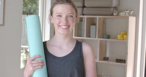 Portrait of happy caucasian fitness teenage girl holding yoga mat in white room, slow motion