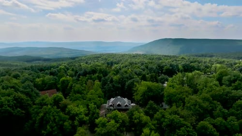 Flying backward against from a house in the forest while hills in the background, on a sunny and clo