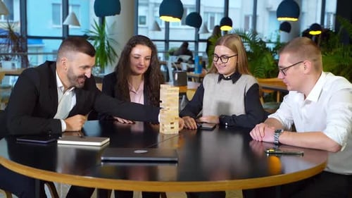 Company employees playing jenga at round table. Man in suit pulls carefully the brick.