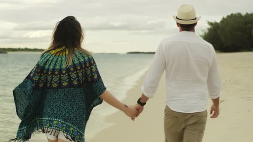 Young Couple Holding Hands and Walking Along the Beach Near the Water