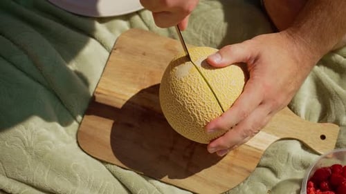Homme tranchant du melon frais sur une planche à découper en bois, préparant soigneusement les fruits pour le pique-nique