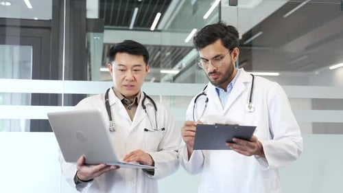 Two doctors in white coats collaborate over a laptop and folder standing in a hospital office.