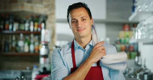 Portrait of a young barman crossing arms and smiling at the camera, behind the bench of a cafe. Co