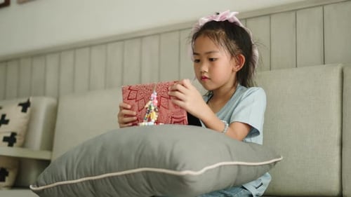 Little Asian Girl with a Bow on Her Head Using Digital Tablet While Sitting Comfortably on Sofa