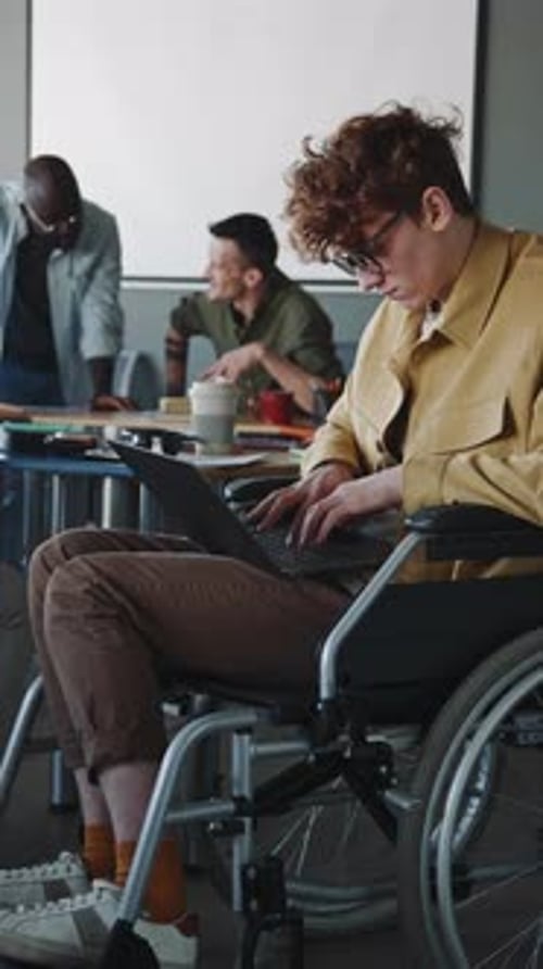 Young Man with Disability Sitting in Wheelchair and Working on Laptop in Office