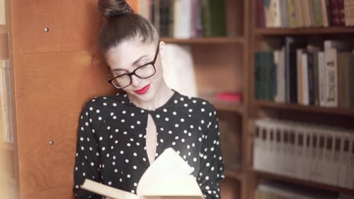 Woman Flicking Through Pages Of A Book While Leaning Against A Shelf In A Library