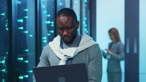 A Man Works on His Laptop in a Server Room Showcasing Tech Skills