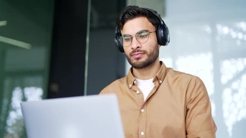 Confident IT specialist developer in wireless headphones works on laptop computer sitting
