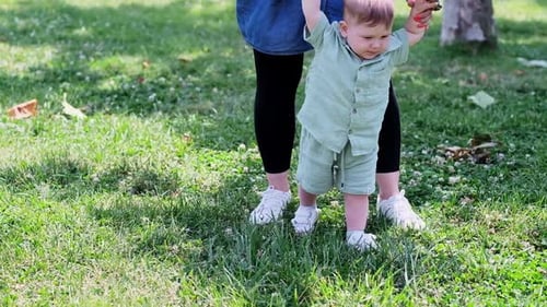 Cute Baby Boy Learning to Walk with Careful Mother Help on Lush Lawn Grass in Spring Park